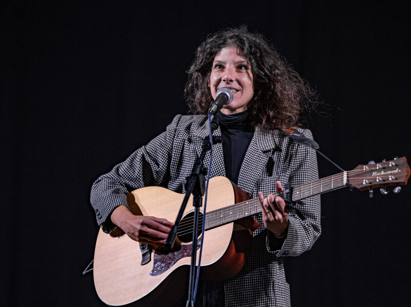 Isabella Del Fagio che suona la chitarra e canta al microfono su un palco, illuminata da una luce frontale su sfondo scuro.