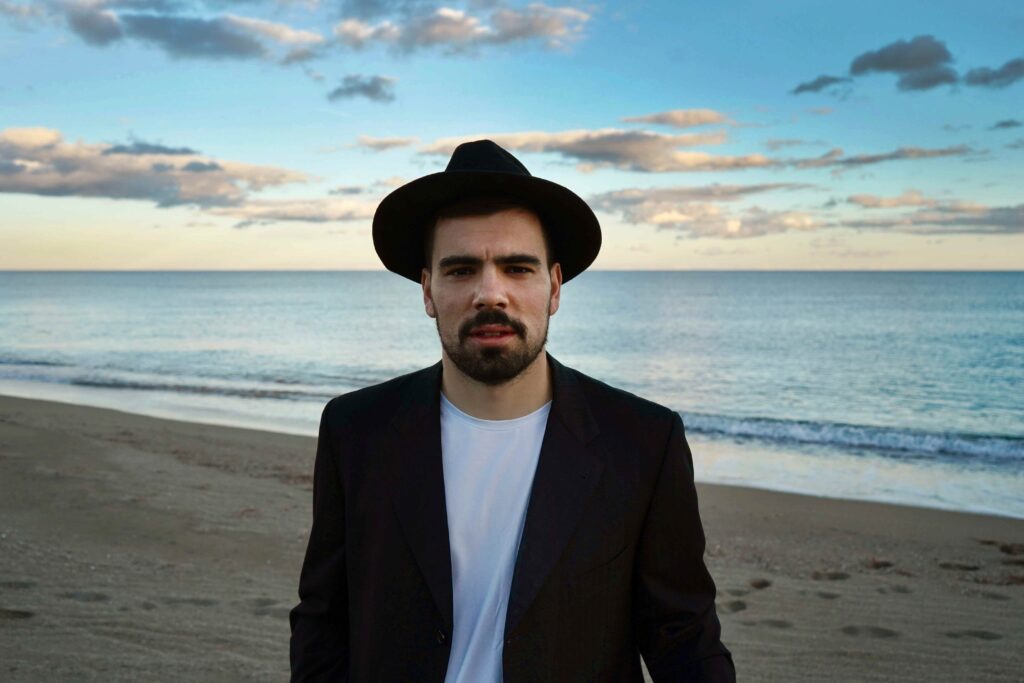 Tommy Mauri in primo piano sulla spiaggia al tramonto, indossa un cappello nero e una giacca scura, con il mare sullo sfondo.