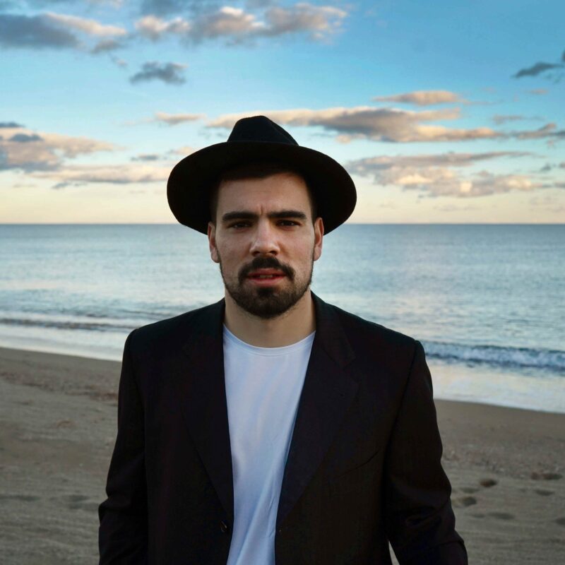 Tommy Mauri in primo piano sulla spiaggia al tramonto, indossa un cappello nero e una giacca scura, con il mare sullo sfondo.