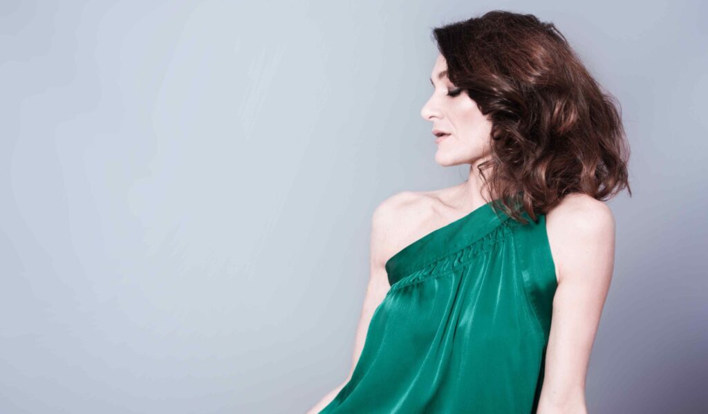 Cristina Zavalloni in posa su fondo neutro, indossa un abito verde elegante monospalla, seduta di profilo con lo sguardo rivolto verso il basso.